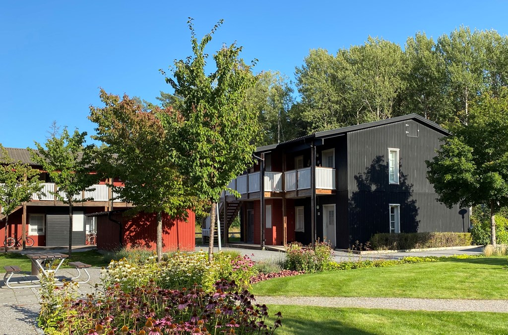 Svarta och röda trähus med vita loftgångar med gräsmatta, grusad gångväg, blomrabatter och en sittg