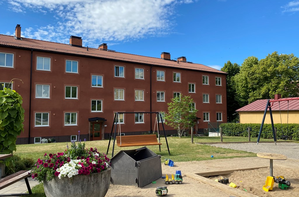 Brunfärgat trevånings flerbostadsshus med bruna balkonger med en gräsmatta och en lekplats, blommor
