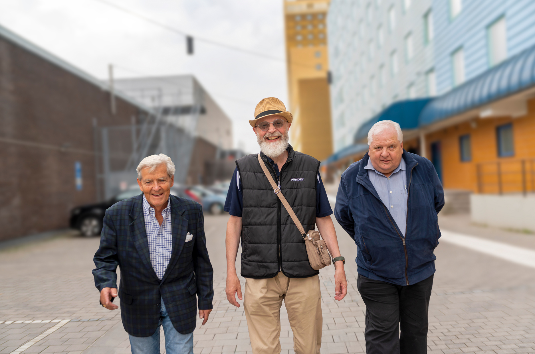 Tre äldre män går bredvid varandra med hus i bakgrunden.