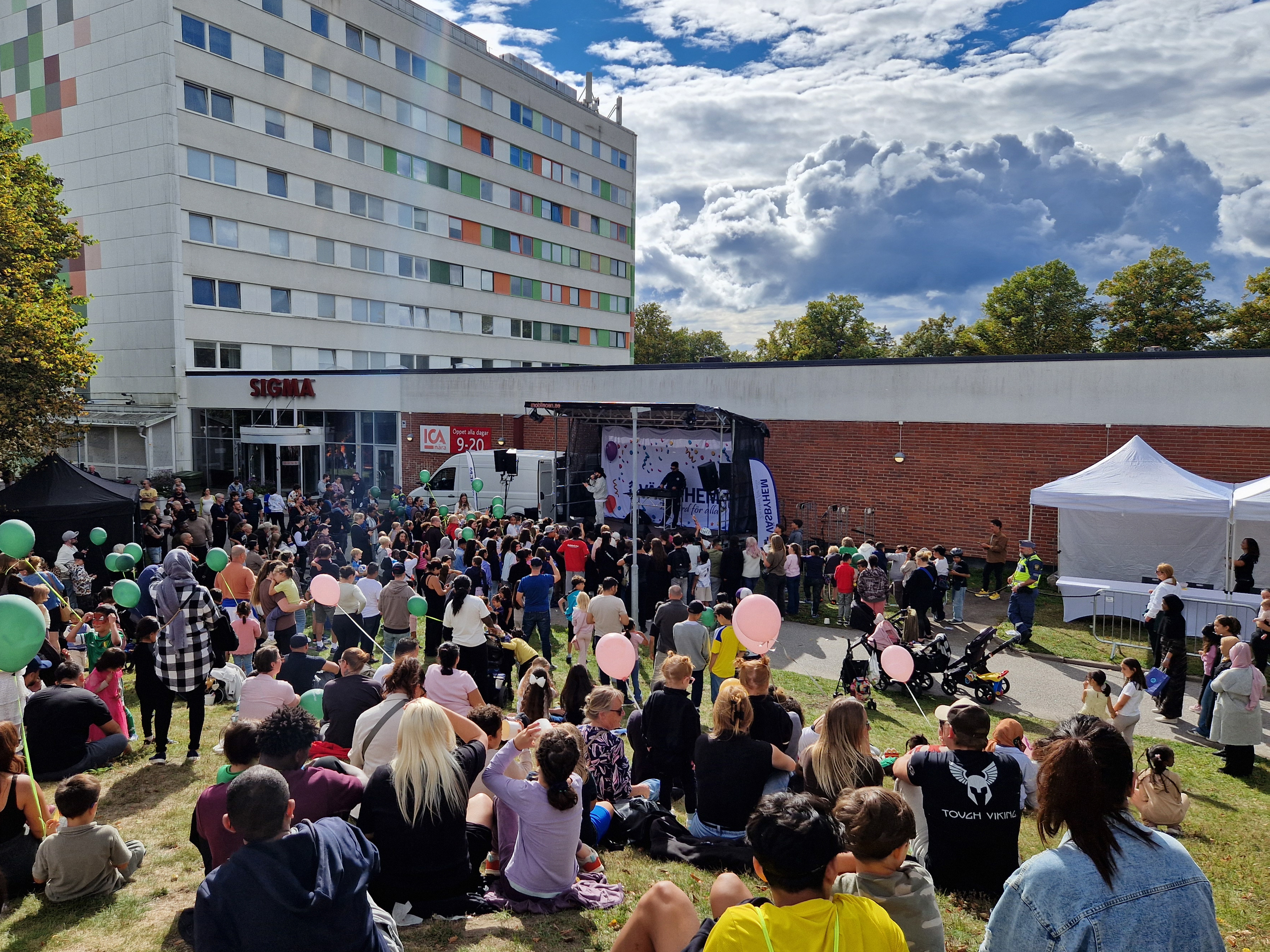 Människor framför scenen utomhus med byggnad i bakgrunden.